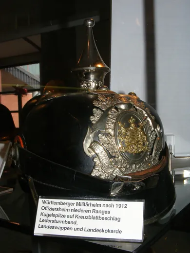 Casque noir d'officier subalterne wurtembergeois postérieur à 1912, en vitrine : boule sur base cruciforme, armoiries du Wurtemberg, cocarde de l'État et jugulaire de cuir