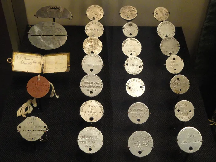 Vitrine de plaques d'identité de plusieurs nations : disques ronds américains en aluminium, disque de fibre rouge britannique, plaque française sécable de 1918 et livret d'identité